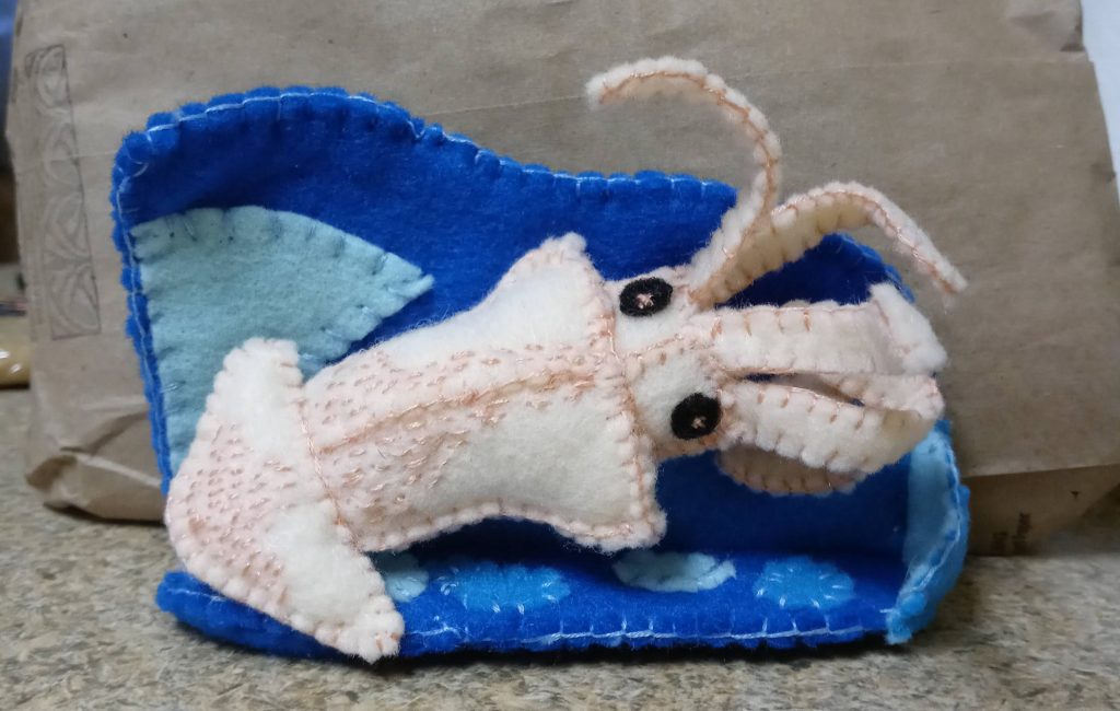 a soft sculpture of a light pink squid in front of an embroidered background of various shades of blue made to imitate a wave.