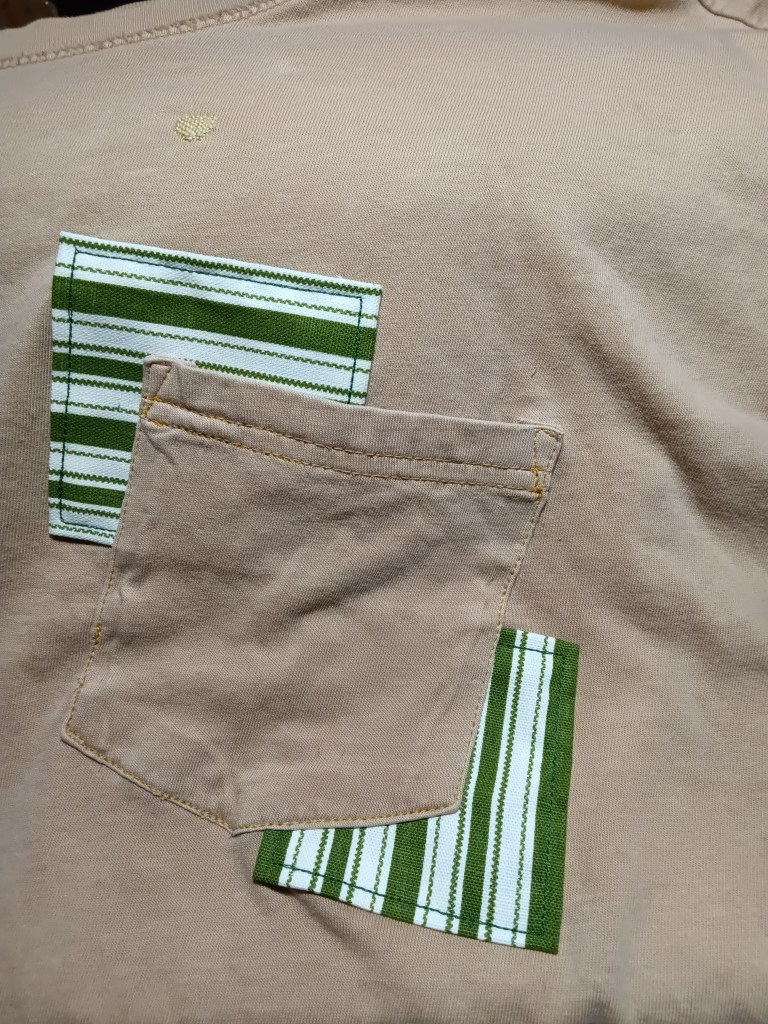 An orange shirt with an orange pocket which is laid on top of two patches of green and white stripes.