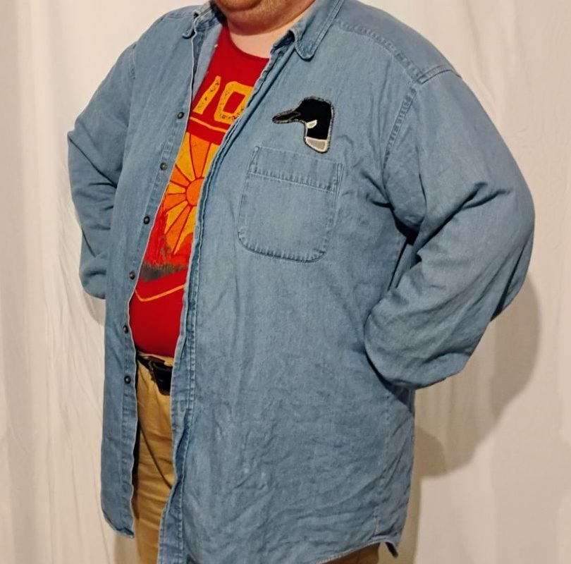 A person standing with their hands on their hips. They are wearing a light blue denim shirt over a red shirt. The denim shirt has a pocket and above the pocket is a patch of a loon head.