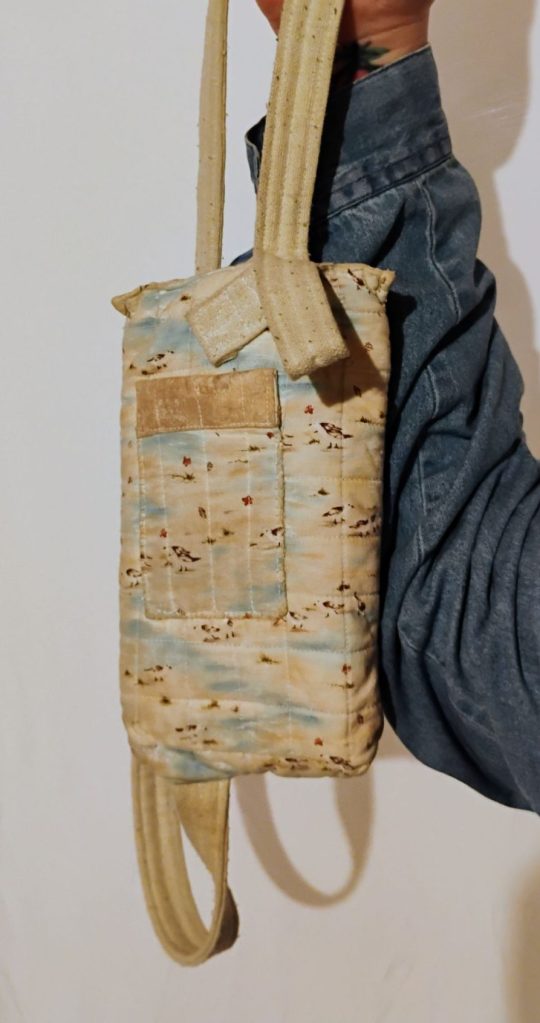 A hand holding out a rectangular bag, showing the back of it. The strap is off-white and the bag is a mix of tan and blue and white with brown spots. The back of the bag features a rectangular patch pocket made of the same fabric with a sandy colored stripe on top.