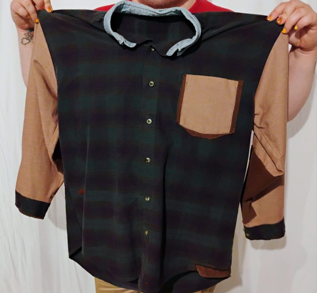 A person holding a long sleeve button up shirt. The body of the shirt is blue and green plaid. The collar is light blue. The sleeves and breast pocket are a pale red. There are various brown patches on the shirt.