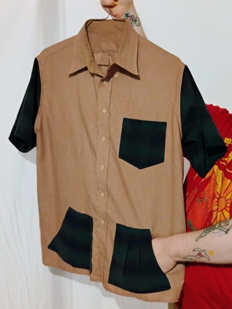 A pale red button up t-shirt. It has a green and blue plaid breast pocket, sleeves, and bottom pockets. The person holding the shirt has their hand in the pocket.