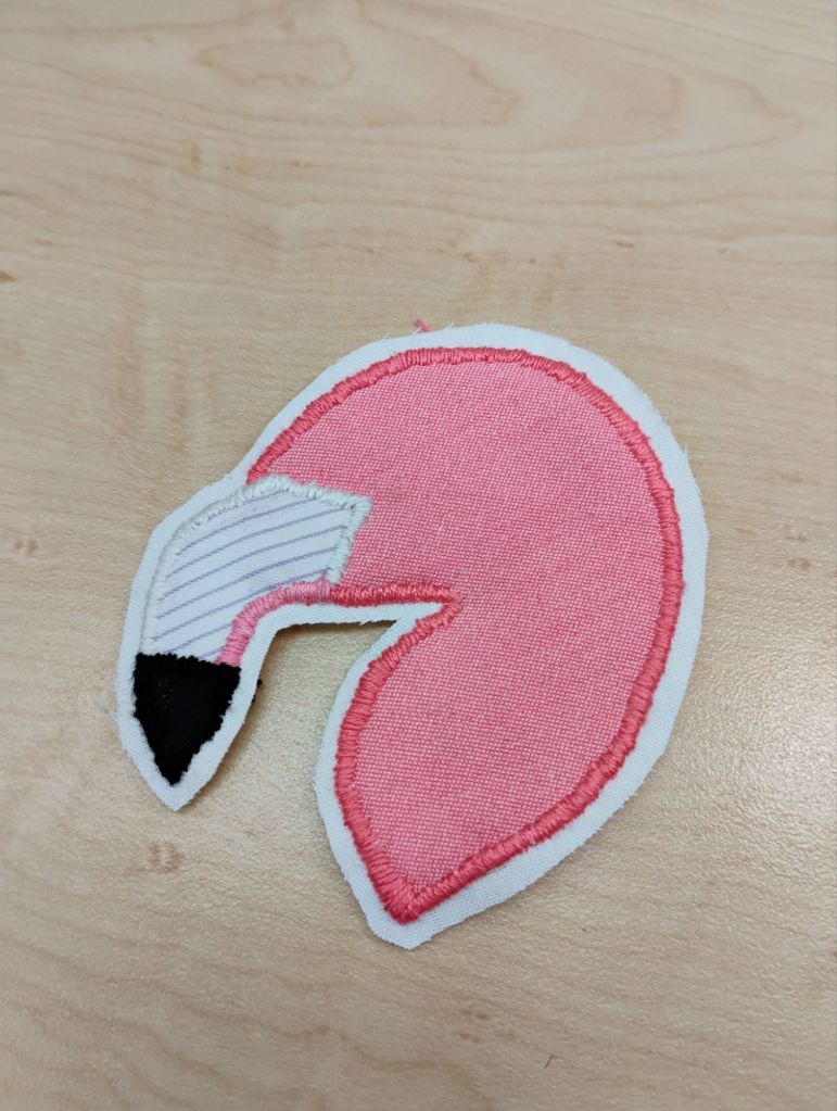 An embroidered patch of the head of a flamingo. it is pink with a white hook beak that has a black tip.