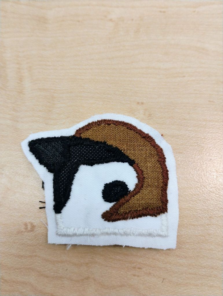 An embroidered patch of the head of an eurasian tree sparrow. It has a white head with a black mask, beak, and cheek spot, as well as a brown bit on the top of its head