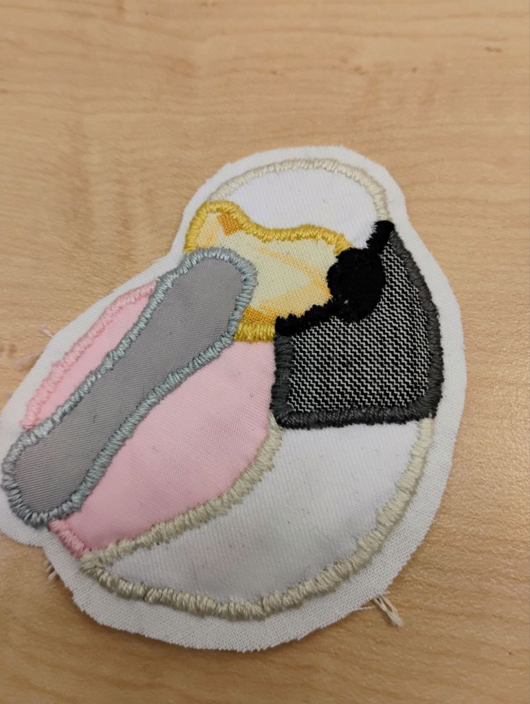 An embroidered patch of the head of a roseate spoonbill. it has a pink circle background, a yellow mask, a grey neck stripe, and a light grey long bill.