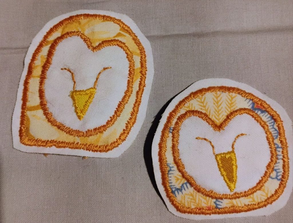 Two embroidered patches of the head of a barn owl. They have white faces with yellow heads, a long beak, and lines resembling closed eyes.